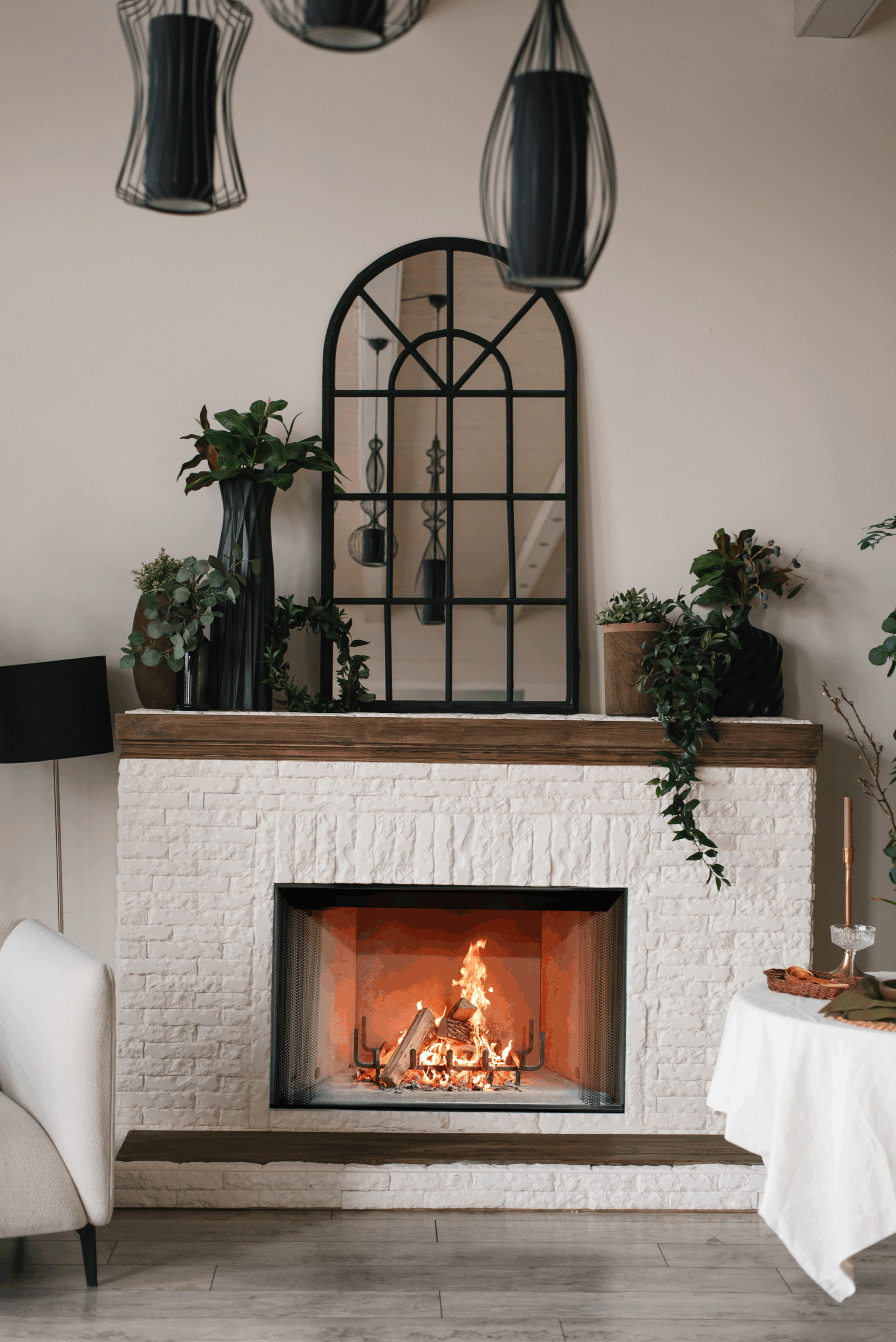 A modern indoor fireplace scene with a white brick fireplace insert, glowing fire, dark wood mantel, hanging pendant lights, a black arched mirror, and plants in a styled room.
