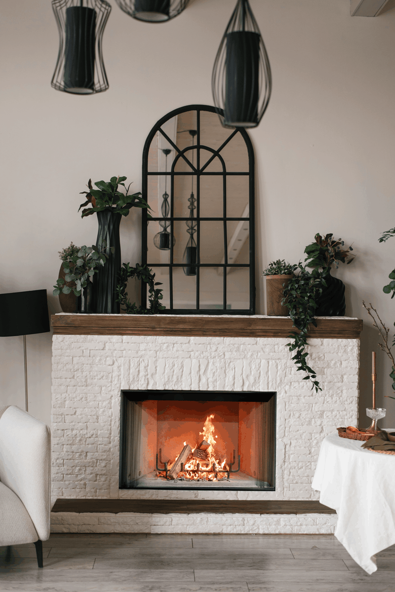 A modern indoor fireplace scene with a white brick fireplace insert, glowing fire, dark wood mantel, hanging pendant lights, a black arched mirror, and plants in a styled room.